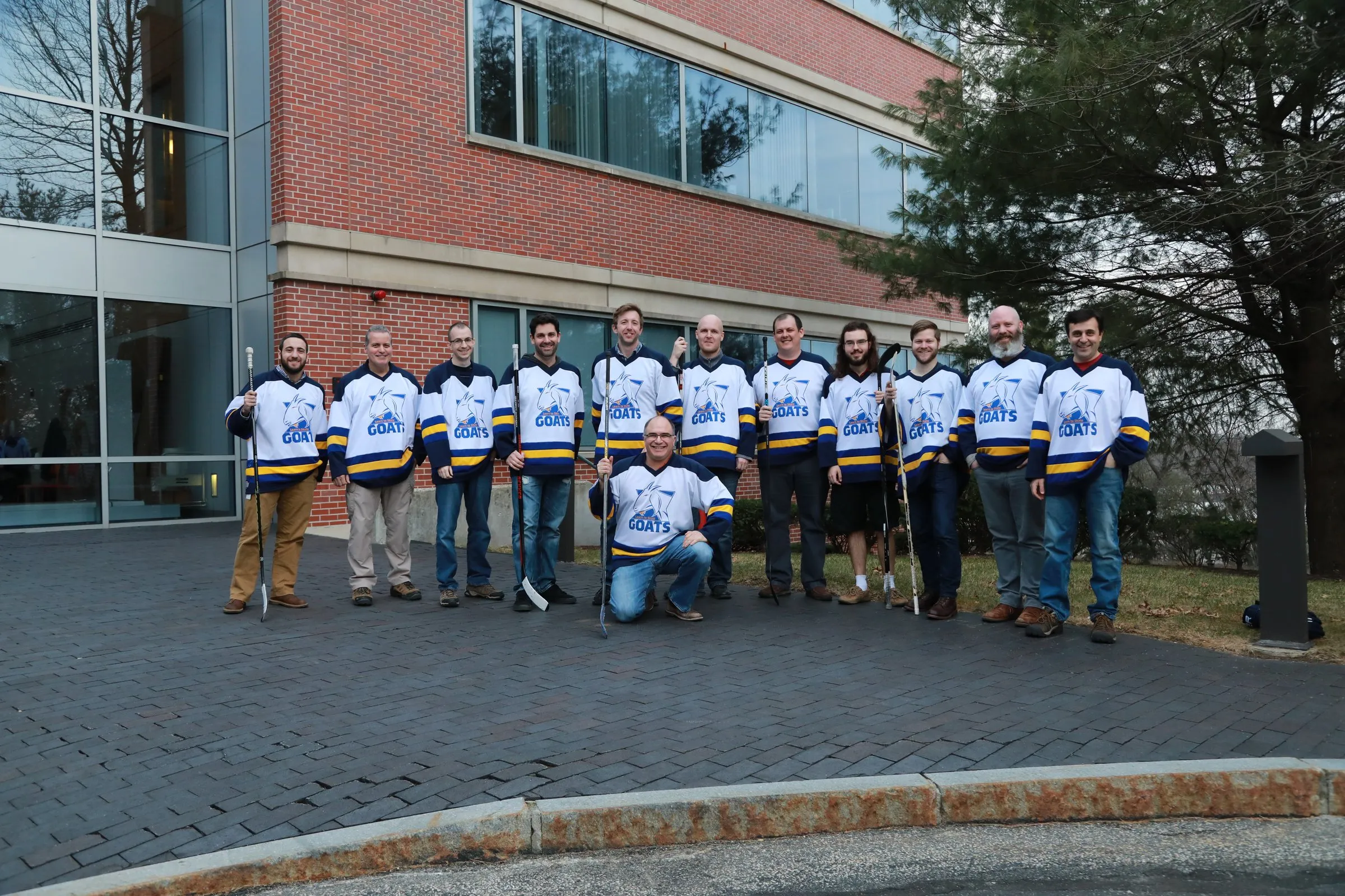 The Goats team in their jerseys, holding hockey sticks in front of an office building